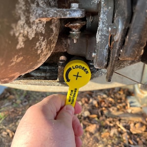 Puede incluir: Una herramienta amarilla con la palabra "LOOSEN" y un s&iacute;mbolo de flecha se sostiene cerca de un componente met&aacute;lico. La herramienta tambi&eacute;n tiene el texto "NOT FOR TIGHTENING". El fondo muestra partes de un veh&iacute;culo.