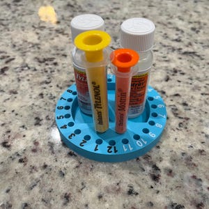May include: A blue plastic medicine dispenser with a circular base and 12 numbered slots. Two clear plastic bottles with yellow and orange droppers are in the dispenser. The bottles are labeled "Infants' TYLENOL" and "Infants' Motrin".