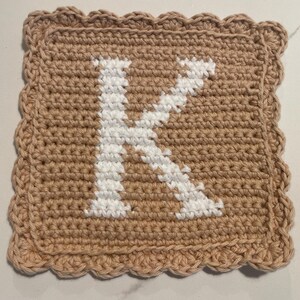 May include: A square, crocheted item in a light brown color, featuring a white letter "K" in the center. The edges are finished with a scalloped border. This handmade item is likely a decorative piece or part of a larger craft project.
