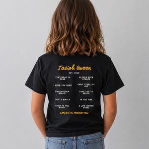 May include: A black t-shirt with the text "Jasiah Queen EST. 2020" in gold script. Below the text are song titles in a vertical list. The shirt is worn by a person with long brown hair and blue jeans.