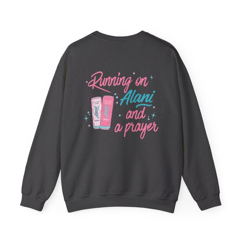 May include: Dark gray sweatshirt with the text "Running on Alani and a prayer" in pink and teal. The design includes illustrations of two Alani Nu energy drink cans. A casual, comfortable top.