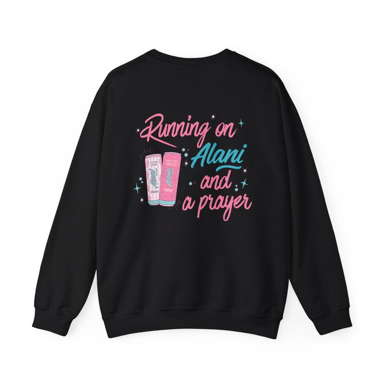 May include: Black sweatshirt with the text "Running on Alani and a prayer" in pink and teal. The design includes illustrations of two Alani Nu energy drink cans. A casual and comfortable apparel item.