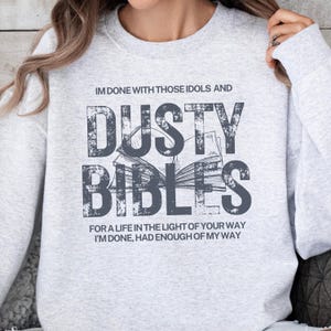 May include: Light gray sweatshirt with the text "IM DONE WITH THOSE IDOLS AND DUSTY BIBLES" in a distressed font. Below the text is an open book graphic. The bottom of the sweatshirt reads "FOR A LIFE IN THE LIGHT OF YOUR WAY I'M DONE, HAD ENOUGH OF MY WAY."
