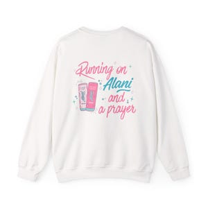 May include: White sweatshirt with the phrase "Running on Alani and a prayer" in pink and blue script. Two beverage cans are depicted in pink and blue. The sweatshirt is a casual, comfortable style.