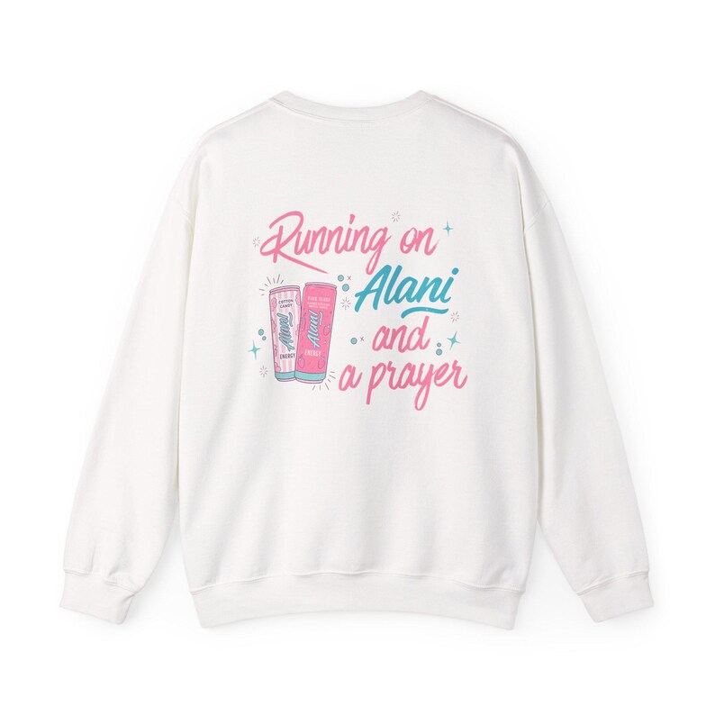 May include: White sweatshirt with the phrase "Running on Alani and a prayer" in pink and blue script. Two beverage cans are depicted in pink and blue. The sweatshirt is a casual, comfortable style.
