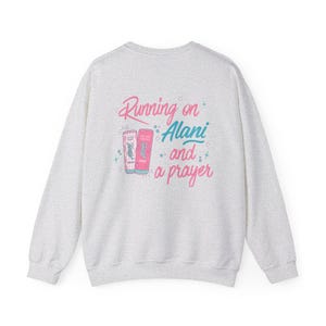 May include: A light gray sweatshirt with the phrase "Running on Alani and a prayer" in pink and teal lettering. The design includes illustrations of two Alani Nu energy drink cans.