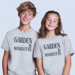 May include: Two people wearing light gray t-shirts with the text "GARDEN IN MANHATTAN" in dark blue. The text is above a skyline graphic. The t-shirts are short-sleeved and appear to be made of a soft material.