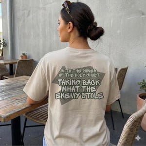 May include: A beige t-shirt with the text "WITH THE POWER OF THE HOLY GHOST TAKING BACK WHAT THE ENEMY STOLE" printed on the back in a gray diamond shape. The person is sitting at a wooden table.