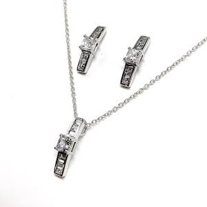May include: A silver necklace and earrings set with a square-shaped clear crystal pendant and matching earrings. The pendant and earrings are accented with smaller clear crystals.