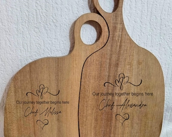 Pode incluir: Duas tábuas de corte de madeira com acabamento castanho claro. Cada tábua tem uma pega com um furo e é gravada com o texto "Our journey together begins here" e o nome de um chef. As tábuas estão lado a lado.
