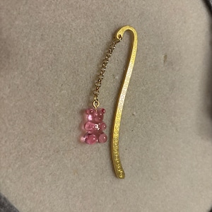 May include: A gold-colored metal bookmark with a curved, S-shaped design featuring a repeating spiral pattern. A thin gold-colored chain is attached to the top, ending in a small, translucent pink gummy bear-shaped charm made of resin.
