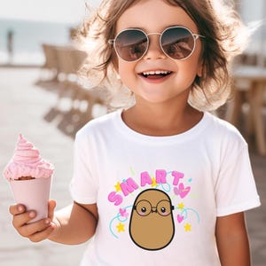May include: A young child wearing a white t-shirt with a cartoon potato design and the word "SMART" in pink letters. The child is smiling and wearing sunglasses, holding a pink ice cream cone.