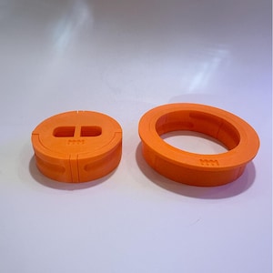 May include: Two orange 3D-printed objects. One is a circular, flat piece with two rectangular cutouts and a series of small lines. The other is a ring-shaped object with a similar series of small lines. Both objects are on a white surface.
