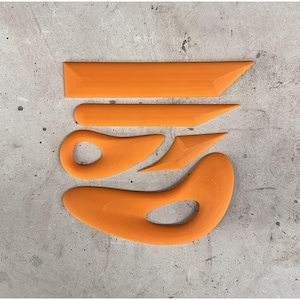 May include: An assortment of orange pottery tools, including a long, flat rib, a pointed rib, a loop tool, a triangle-shaped tool, and a curved tool. The tools are arranged on a gray concrete surface.
