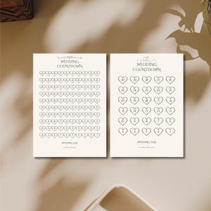May include: Two cream-colored wedding countdown charts. Each chart features a series of numbered hearts. The text "WEDDING COUNTDOWN" is at the top, and "WEDDING DAY" is at the bottom. The charts are designed to track the days until a wedding.