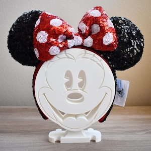 Op de afbeelding: Zwart en rood Minnie Mouse orenband met witte stippen. Het hoofdband staat op een witte standaard.