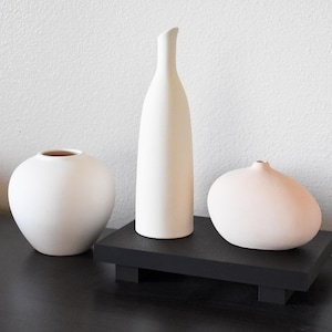 May include: Three white ceramic vases of different shapes and sizes on a black wooden stand. The vases are all simple and modern in design.