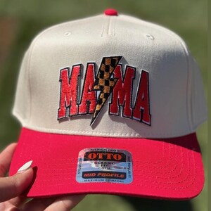 Può includere: Un cappellino da baseball beige e rosso con la scritta "MAMA" a blocchi rossi e un fulmine a scacchi. Il cappellino ha una visiera rossa e un bottone rosso sulla parte superiore. L'etichetta indica "MID PROFILE".