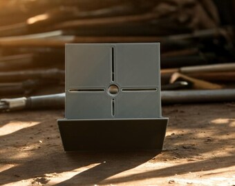 May include: A grey, geometric object with a square top and a rectangular base. The top has a cross-shaped cutout with four rectangular sections and a circular centre. The object is made of a matte material and sits on a surface.