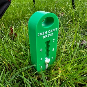 May include: Green plastic golf scorekeeper with the text "Josh Can't Drive" and numbers 2 to 7. A white golf tee is inserted into the scorekeeper.