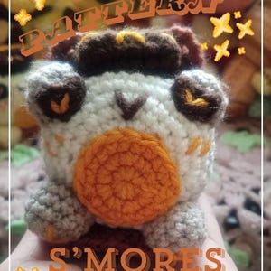 May include: Crochet pattern for a s'mores frog amigurumi. The pattern is available as a PDF download. The frog is made with brown, white and orange yarn. The frog has a brown hat and a large orange s'more on its belly.