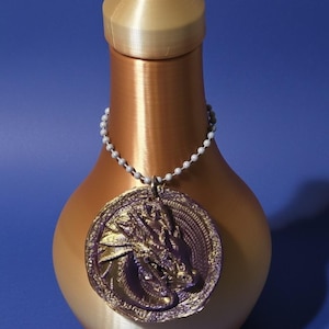 May include: A decorative, metallic-colored bottle with a matching lid. A detailed dragon medallion hangs from a beaded chain around the bottle's neck. The bottle has a gradient finish, transitioning from gold to copper.