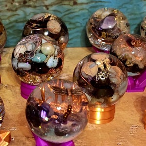 May include: A collection of spherical resin paperweights containing embedded natural stones, metal wire coils, and metallic charms including a dragonfly, a fish, and a bow. Each clear orb is supported by a small, solid-colored plastic stand.