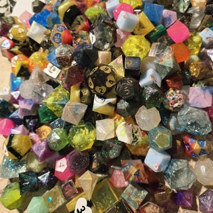 May include: A colorful assortment of polyhedral dice in various shapes and sizes. The dice are made of resin and feature a variety of colors, including pink, blue, green, yellow, and black. Some dice have numbers, while others have decorative elements.