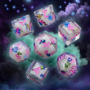Puede incluir: Juego de siete dados de resina transparente con números rosas y diseños florales y de mariposas. Los dados tienen un efecto brillante e iridiscente y están colocados sobre un fondo oscuro y onírico con mariposas y flores flotantes.