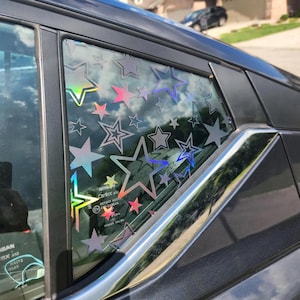 May include: A car window with a holographic star pattern applied. The stars are various sizes and colors, including pink, silver, and iridescent. The window is part of a dark gray car, with a shiny chrome door handle visible.