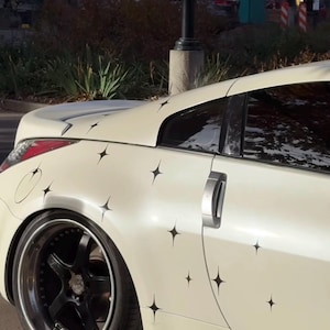 Puede incluir: Coche deportivo color crema con calcomanías de estrellas negras. El coche tiene llantas negras y un tirador de puerta plateado. El piloto trasero es rojo. El coche está aparcado afuera en un día soleado.