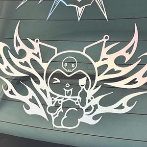 May include: White vinyl sticker of a cartoon character with a skull on its head, winking, sticking its tongue out, and holding a paintbrush. The character is surrounded by a tribal-style design.