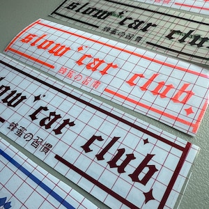 Puede incluir: Cuatro pegatinas de vinilo con el texto "slow car club" en una fuente gótica. Las pegatinas están sobre un fondo de cuadrícula blanca. El texto está en negro, rojo, granate y azul.