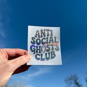 May include: A white sticker with the text "ANTI SOCIAL GHOSTS CLUB" in a holographic, glittery font. The sticker features two small ghost illustrations. The background is a clear blue sky.