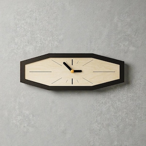 Puede incluir: Un reloj de pared moderno con un marco geométrico negro. La esfera del reloj es de madera clara con líneas negras que marcan las horas y manecillas negras con un punto amarillo. El reloj está montado en una pared gris.
