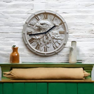 Pode incluir: Um relógio de parede branco redondo com algarismos romanos e um design de águia esculpida. O relógio está pendurado em uma parede de tijolo branco acima de uma prateleira bege com um vaso marrom e um vaso branco.