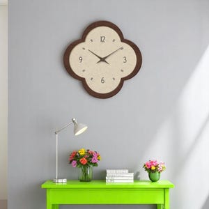 Op de afbeelding: Een bruine houten klok met een witte wijzerplaat en zwarte cijfers. De klok is in de vorm van een klavertjevier en hangt aan een grijze muur. Er staat een groene tafel met een lamp, boeken en bloemen voor de klok.
