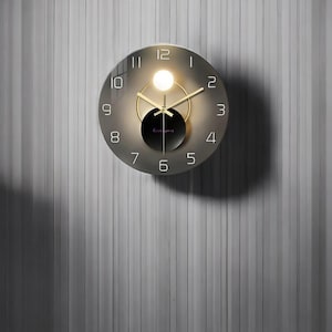 May include: A round, silver-toned wall clock with black numbers and gold hands. The clock has a clear glass face and a black circle in the centre.