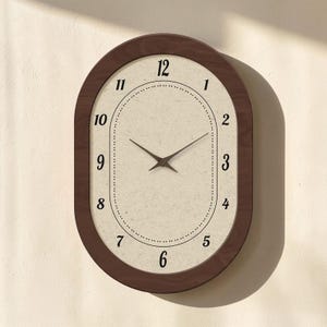 May include: Oval wall clock with a dark brown wooden frame. The clock face is off-white with black numerals and hands. The clock measures approximately 30 cm in height and 23 cm in width.