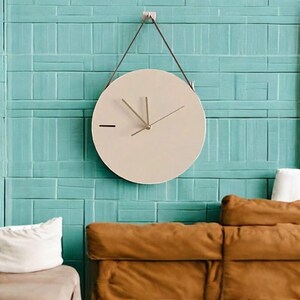 Peut inclure: Une horloge ronde beige avec des aiguilles noires suspendue à un mur turquoise. L'horloge est suspendue par une sangle en cuir marron. Un canapé brun est au premier plan.
