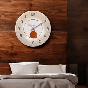 Pode incluir: Um relógio de parede redondo branco com numerais romanos pretos e uma moldura dourada. O mostrador do relógio tem um círculo branco menor com uma moldura dourada e um círculo laranja no centro. Os ponteiros do relógio são dourados.