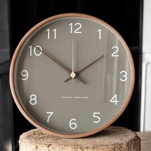 May include: A round, wooden framed clock with a gray face and white numbers. The clock hands are brown and gold. The clock face also includes the text "NEO KOREA ESQUE MJK".