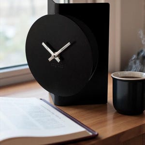 May include: A modern black clock with a circular face and silver hands. The clock is set on a black rectangular base. A black mug of coffee and an open book are on a wooden surface.