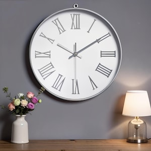 May include: A large, round wall clock with a white face and gray Roman numerals. The clock has a silver-colored frame and hands. The background includes a gray wall, a white vase with flowers, and a small lamp.