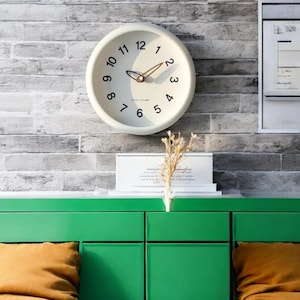 May include: A white round clock with black numbers and hands against a brick wall. The clock is hanging above a green dresser with two drawers. There is a small bouquet of dried flowers on top of the dresser.