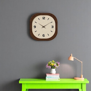 Puede incluir: Un reloj de madera marrón con marco, con una esfera blanca y números negros. El reloj está colgado en una pared gris sobre una mesa verde con un jarrón de flores y una pila de libros.