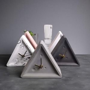 May include: A selection of contemporary triangle-shaped clocks in different colours and patterns. The clocks feature gold-coloured hands and are made of concrete. One has a marble design, others are solid grey, white, and pink.