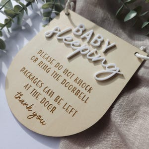 May include: A wooden sign with the words "BABY Sleeping" in white 3D letters. The sign also includes the text "PLEASE DO NOT KNOCK OR RING THE DOORBELL PACKAGES CAN BE LEFT AT THE DOOR thank you". The sign is light brown with a curved bottom.