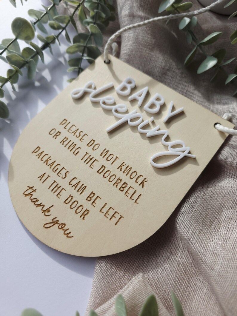 May include: A wooden sign with the words "BABY Sleeping" in white 3D letters. The sign also includes the text "PLEASE DO NOT KNOCK OR RING THE DOORBELL PACKAGES CAN BE LEFT AT THE DOOR thank you". The sign is light brown with a curved bottom.