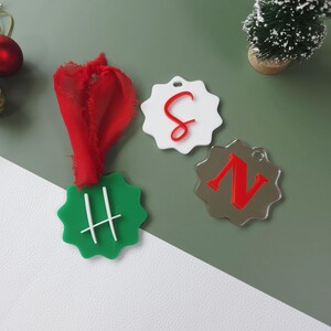 May include: Three decorative tags with scalloped edges. One is green with a white "H", another is white with a red "C", and the third is silver with a red "N". A red ribbon and a small Christmas tree are also visible.